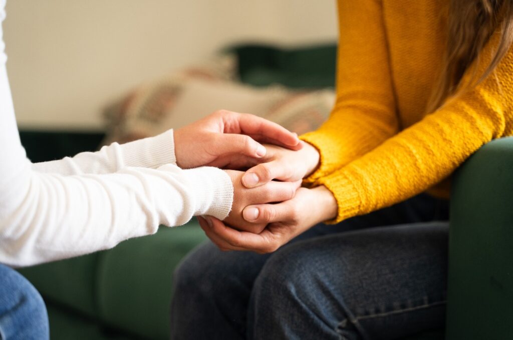 Eine Frau hält die Hand einer anderen Frau während eines herzlichen Gesprächs im Hintergrund des Wohnzimmers, zwei Mädchen halten sich an den Händen als Symbol für weibliche Freundschaft, Trost und Unterstützung zwischen Freunden, Nahaufnahme